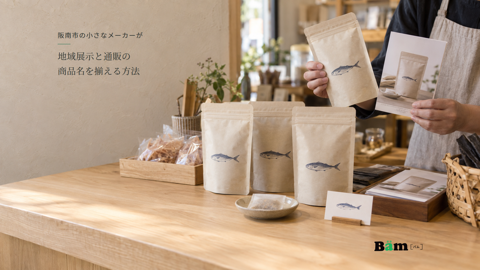 地域の展示台で魚だしの包装と配布資料を照合する手元、阪南市の小さなメーカーが地域展示と通販の商品名を揃える方法の全文タイトル、右下のBämロゴ
