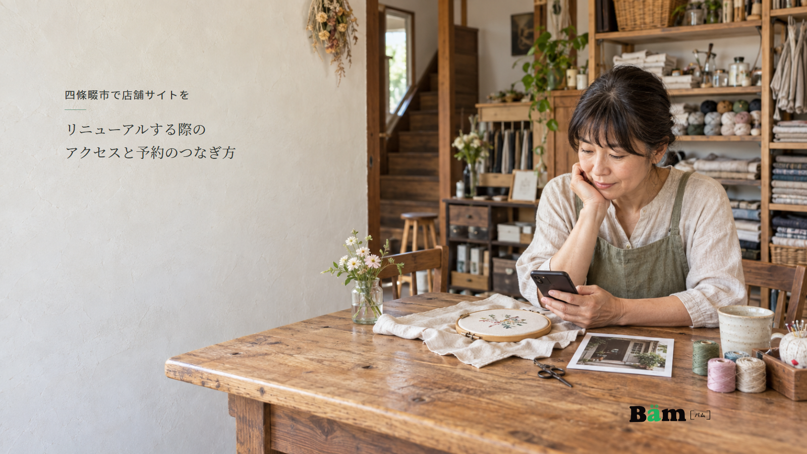 刺繍枠と布が並ぶ手芸店の机でスマートフォンと入口写真を確認する店主、四條畷市で店舗サイトをリニューアルする際のアクセスと予約のつなぎ方の全文タイトル、右下のBämロゴ