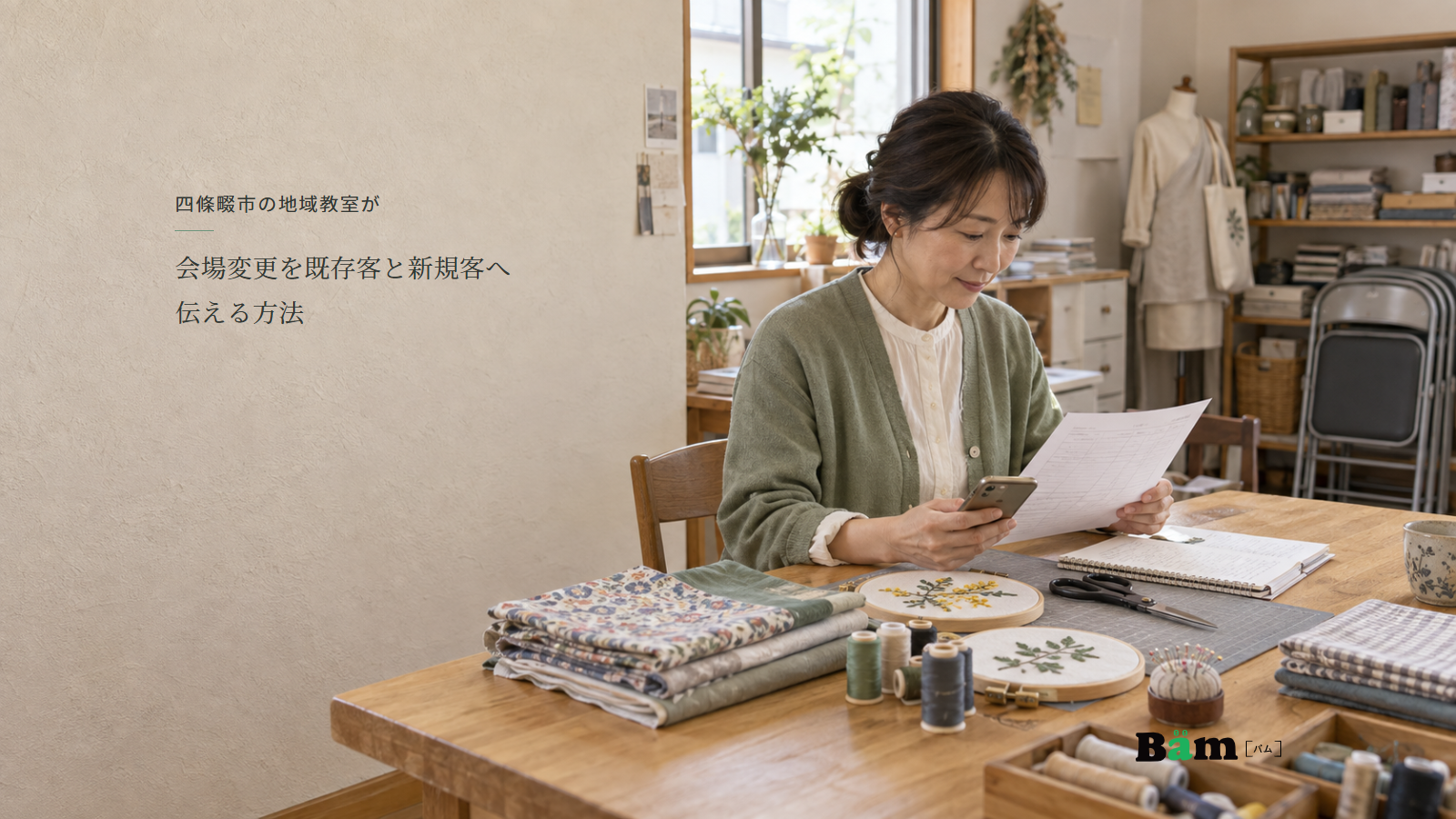 布や刺繍道具が並ぶ教室で講師が開催案内とスマートフォンを確認する場面、四條畷市の地域教室が会場変更を既存客と新規客へ伝える方法の全文タイトル、右下のBämロゴ