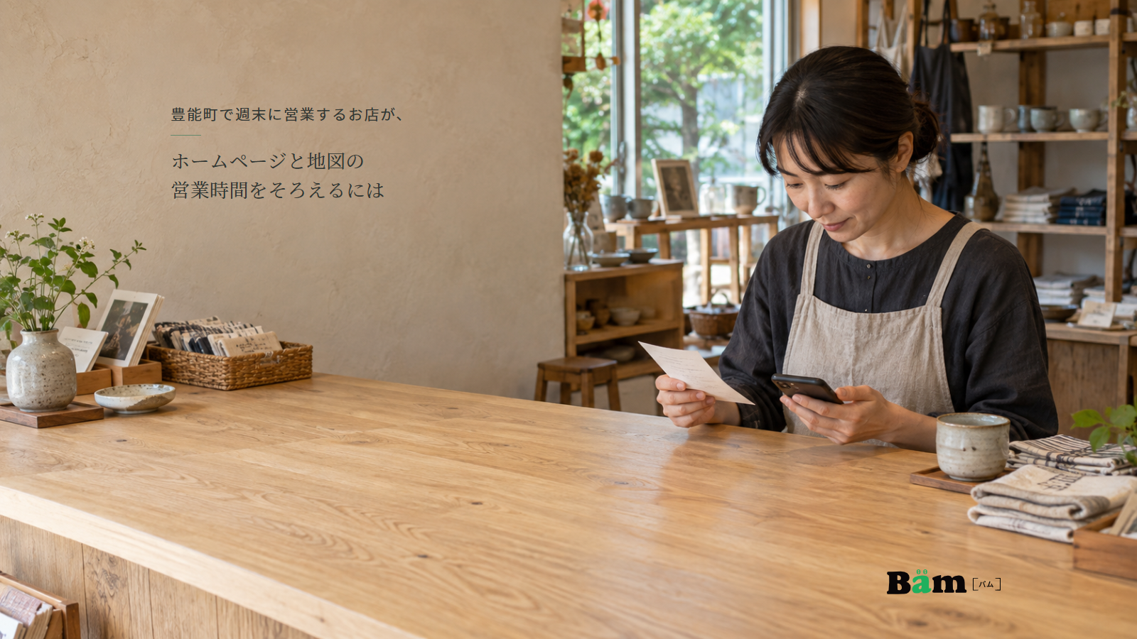 雑貨店の木のカウンターで店主が営業予定の紙とスマートフォンを見比べる様子、豊能町で週末に営業するお店が、ホームページと地図の営業時間をそろえるにはの全文タイトル、右下のBämロゴ